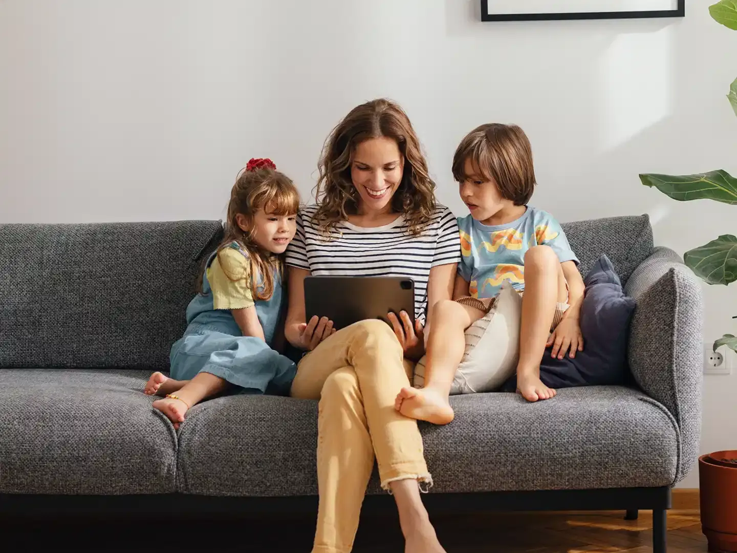 A mother sitting on a couch with her two young children while watching something together on a tablet device.